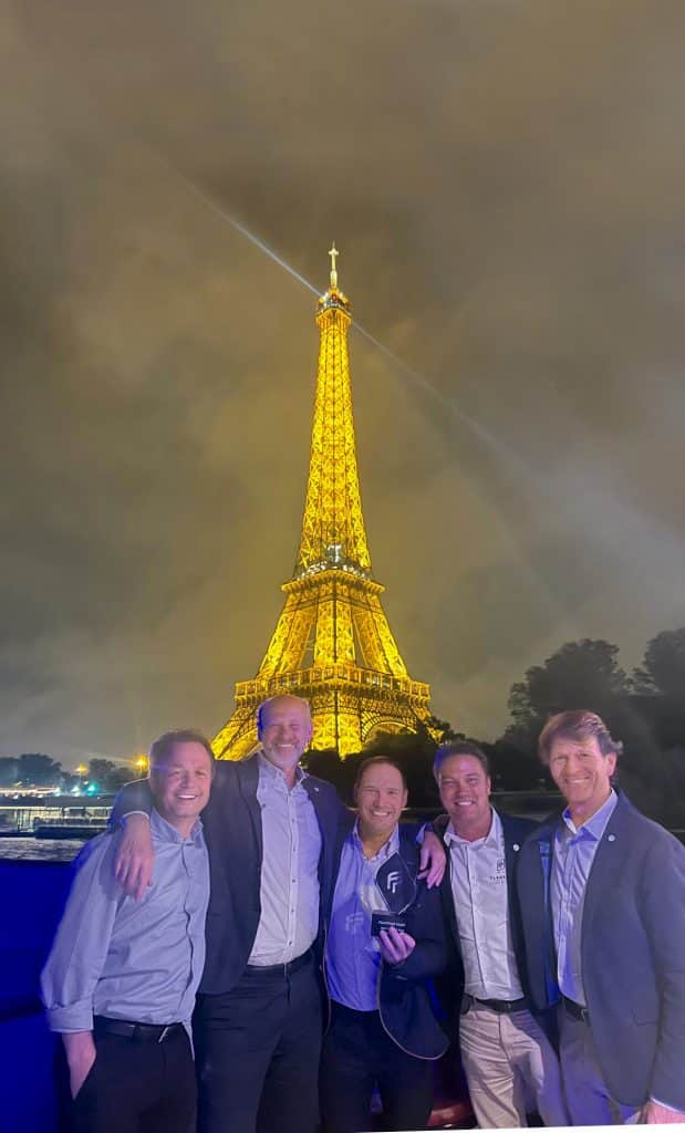 Five men pose together and smile, with the illuminated Eiffel Tower at night in the background. They stand outside in casual business attire and radiate success. The scene looks vibrant, and the lights of the Eiffel Tower create a lively atmosphere reminiscent of a boat dealer's best waxing party.