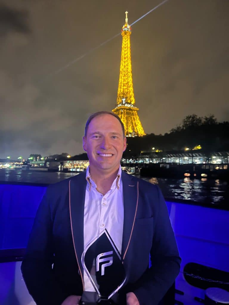 A man in a white shirt and dark blazer holds a trophy in his hand as he stands outside at night. The illuminated Eiffel Tower can be seen in the background, casting its reflection onto the water. The scene suggests that he may be celebrating his success as a top boat dealer for Fountaine Pajot.