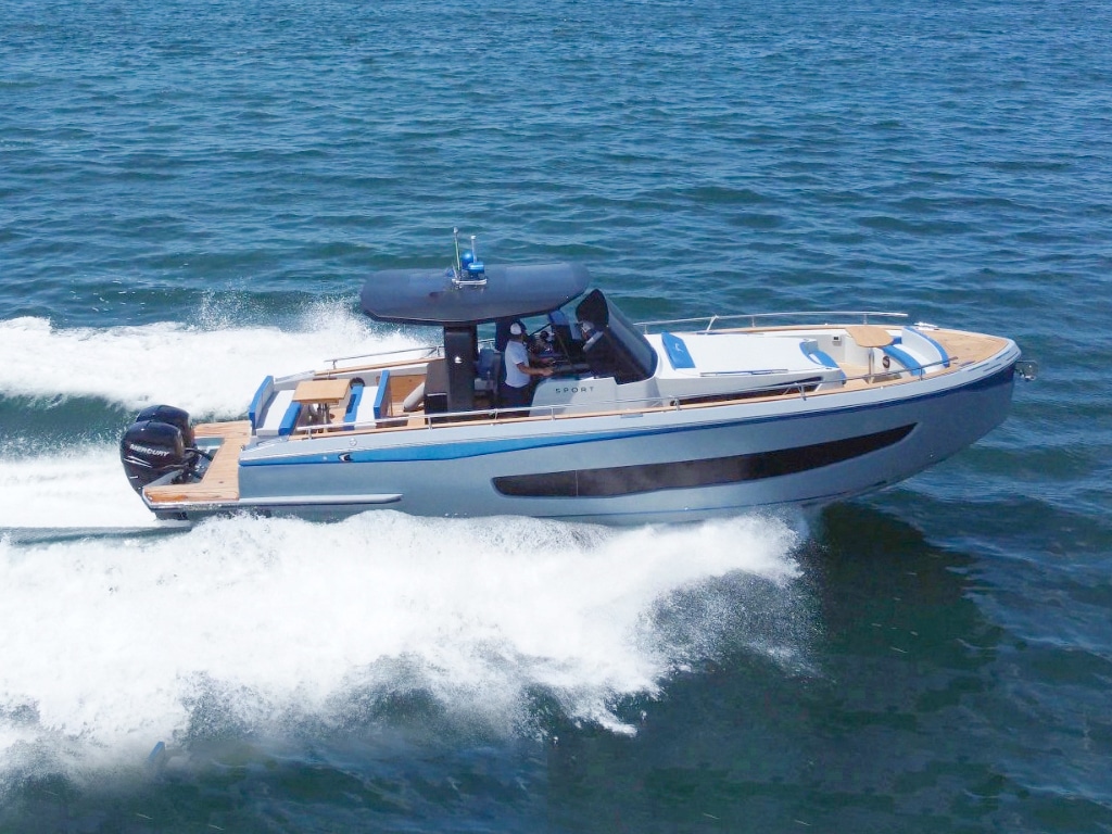 A sleek, modern motorboat races across an expanse of water, leaving a trail of white foam in its wake. The boat has a light gray hull, a dark windshield and a spacious deck area with seating. It is perfect for sightseeing and test drives and has two large outboard motors mounted at the stern.